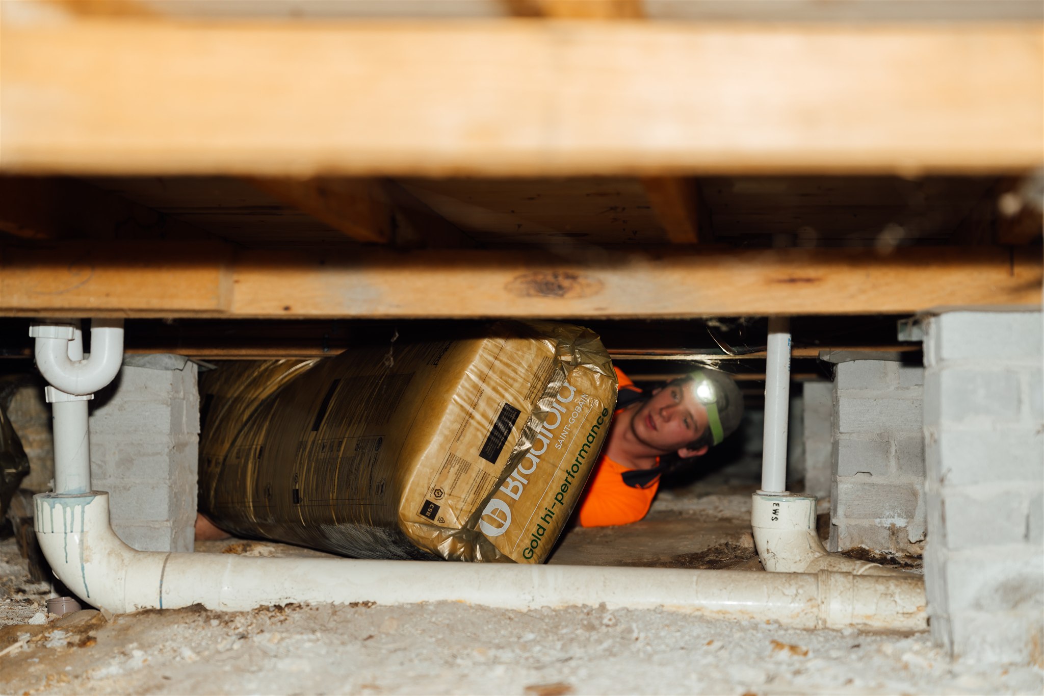 Installer wearing a head torch pushing insulation batts through a subfloor crawl space