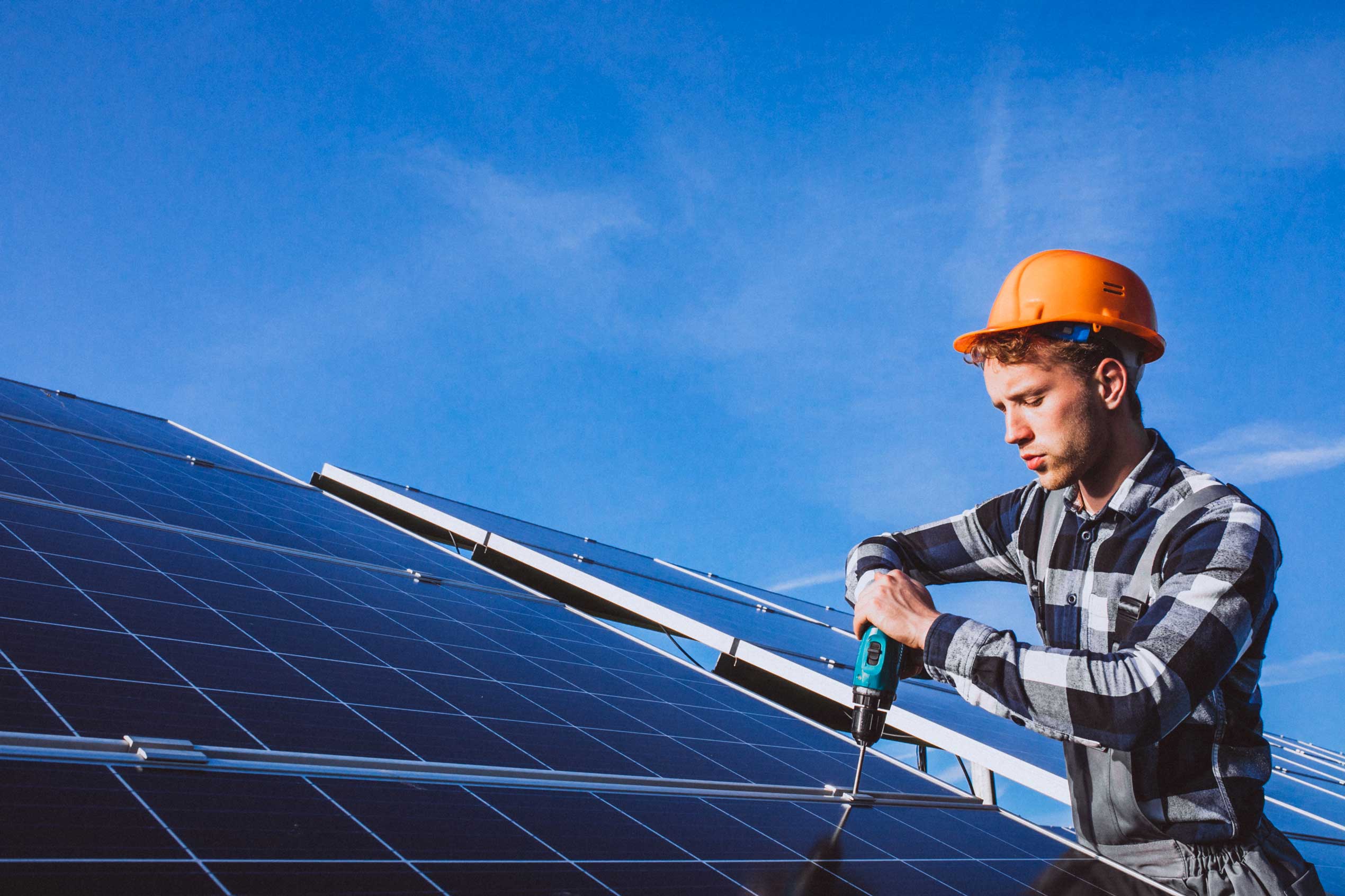 Worker in a hard hat using a drill to install solar panels on a roof against a blue sky