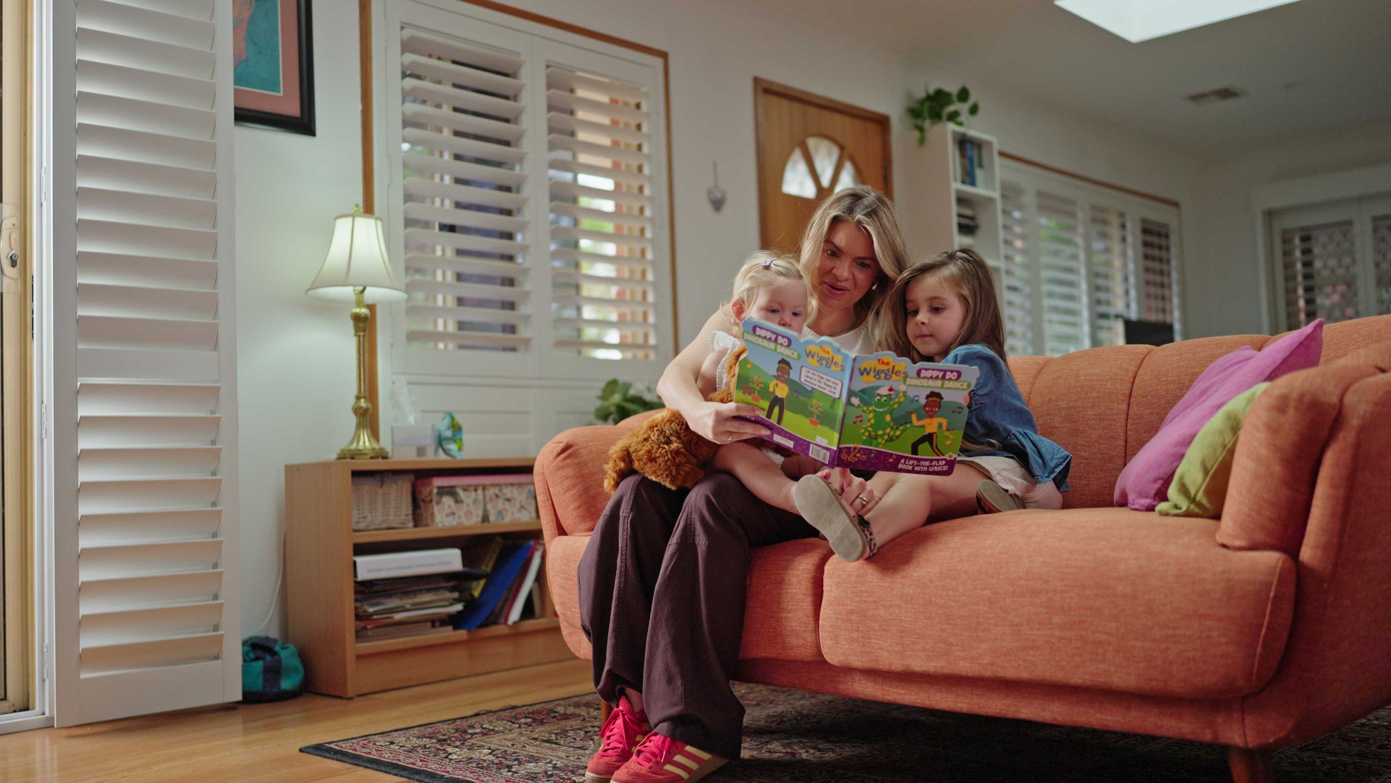 Adult reading a picture book with two young children on a lounge in a sunlit living room