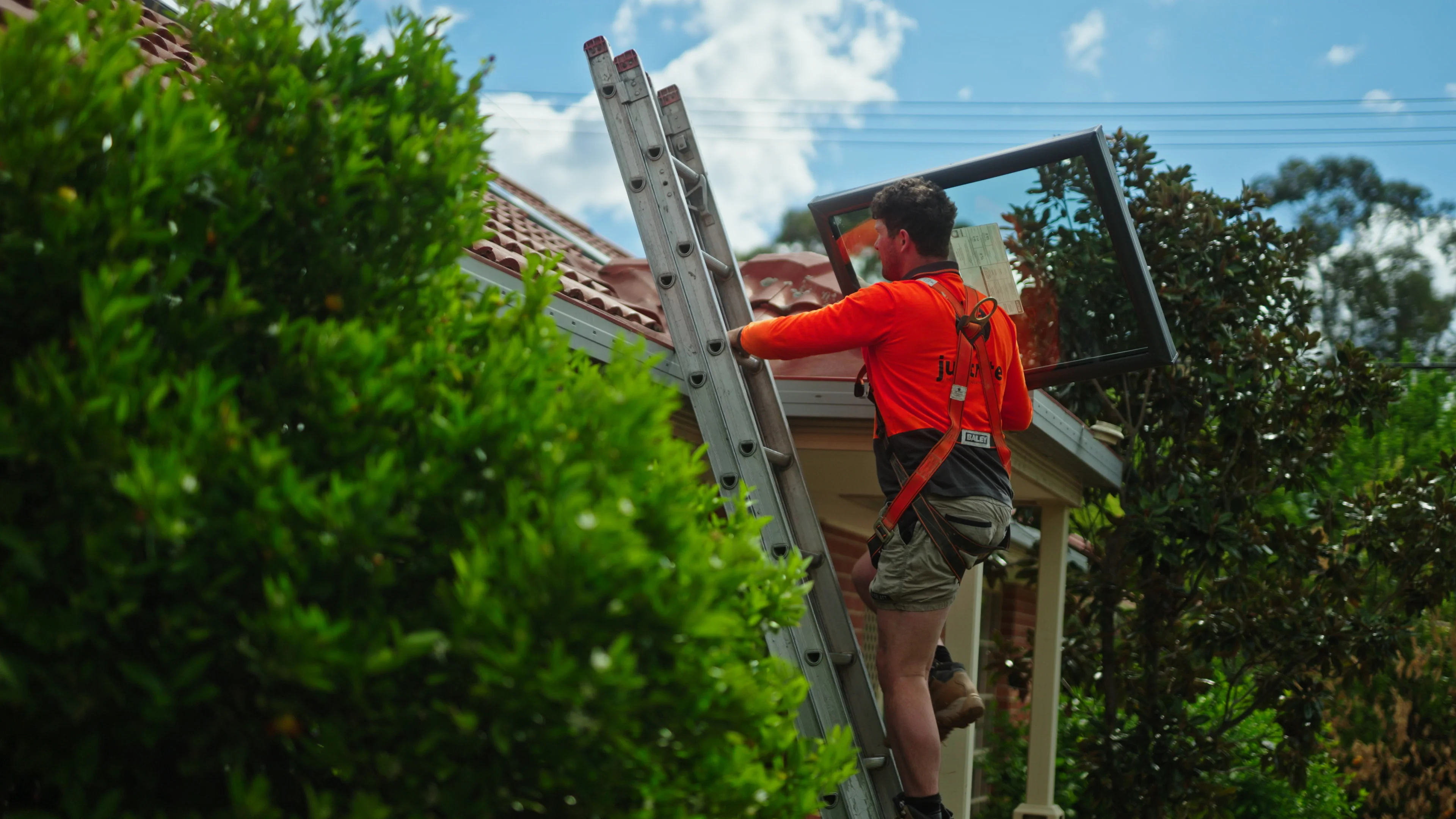 Worker on an extension ladder at the roof edge of a tiled house, working along the gutter line