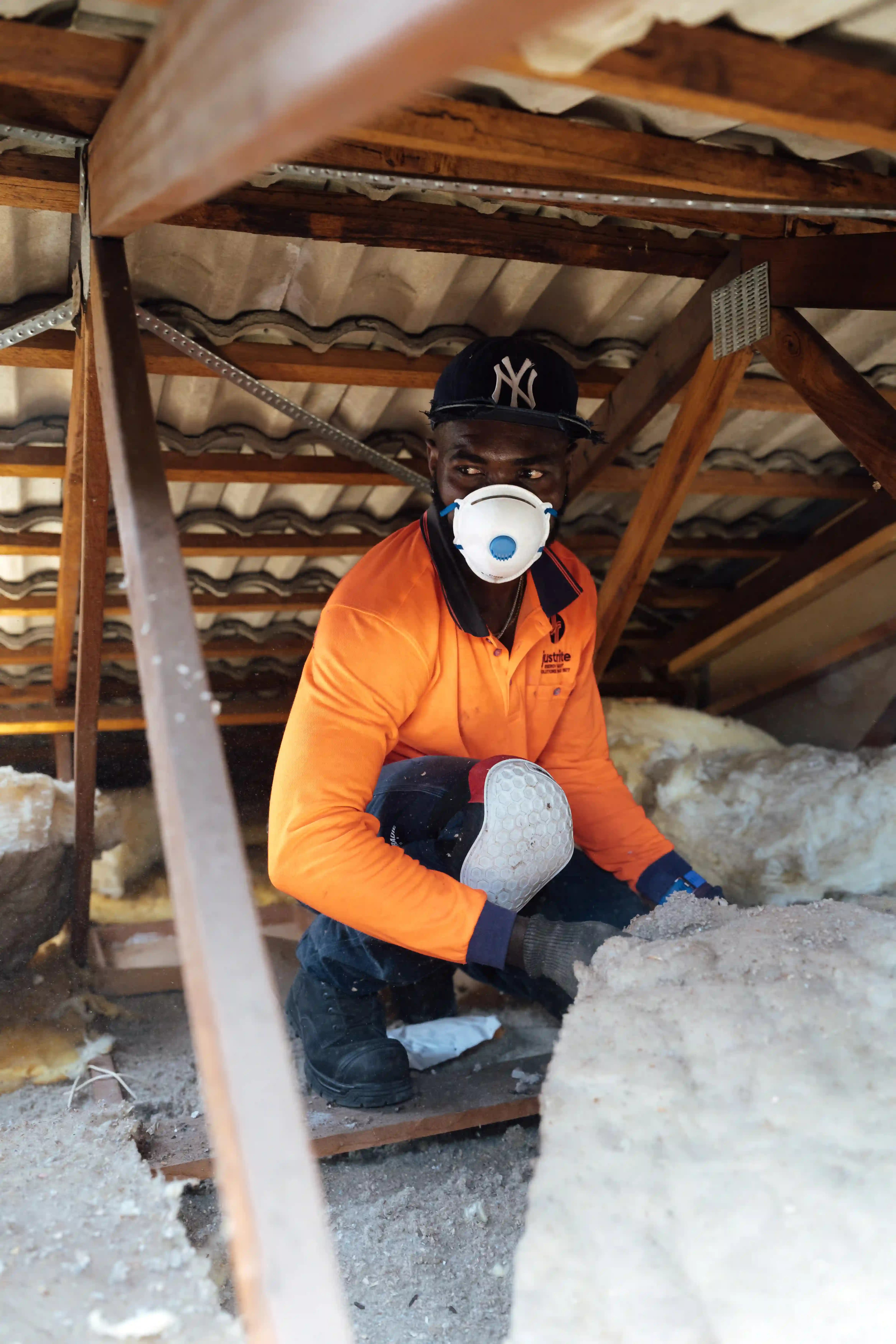 Installer in hi-vis and a dust mask kneeling in a roof cavity while laying ceiling insulation batts