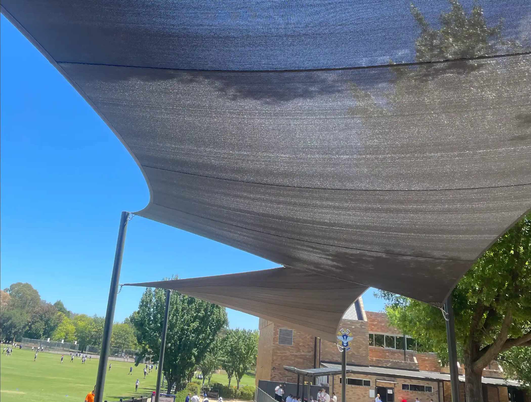 Charcoal shade sails stretched over a paved courtyard beside a grassed oval and brick building