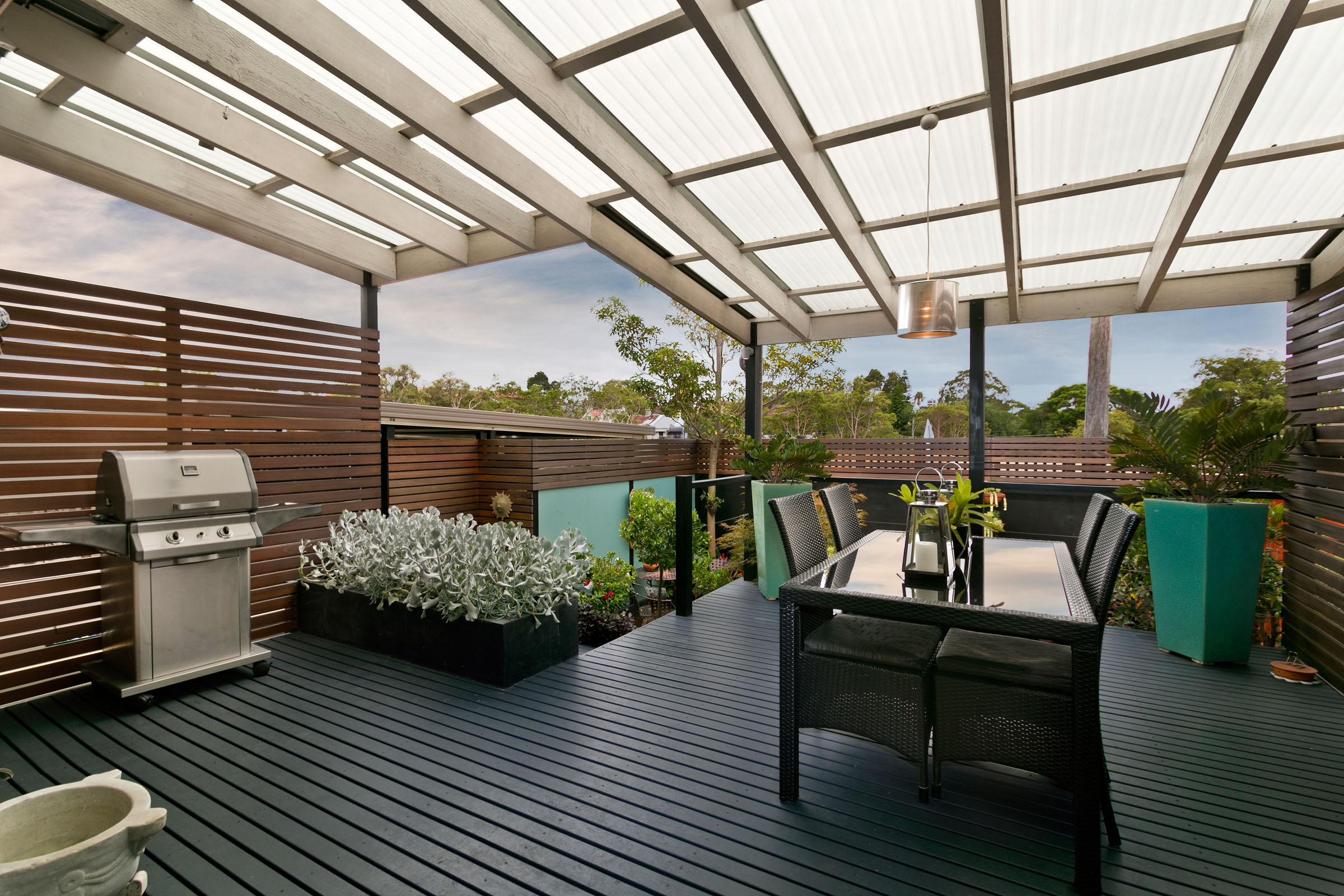 Timber deck with an outdoor dining setting and barbecue under a pergola roofed in clear polycarbonate sheeting
