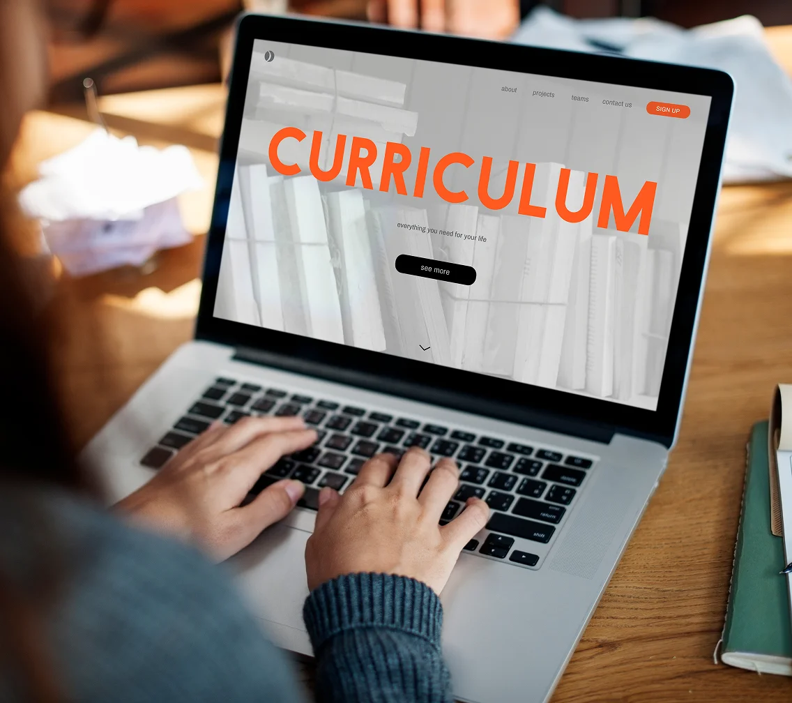 Person typing on laptop showing a webpage with the word CURRICULUM in bold orange letters.