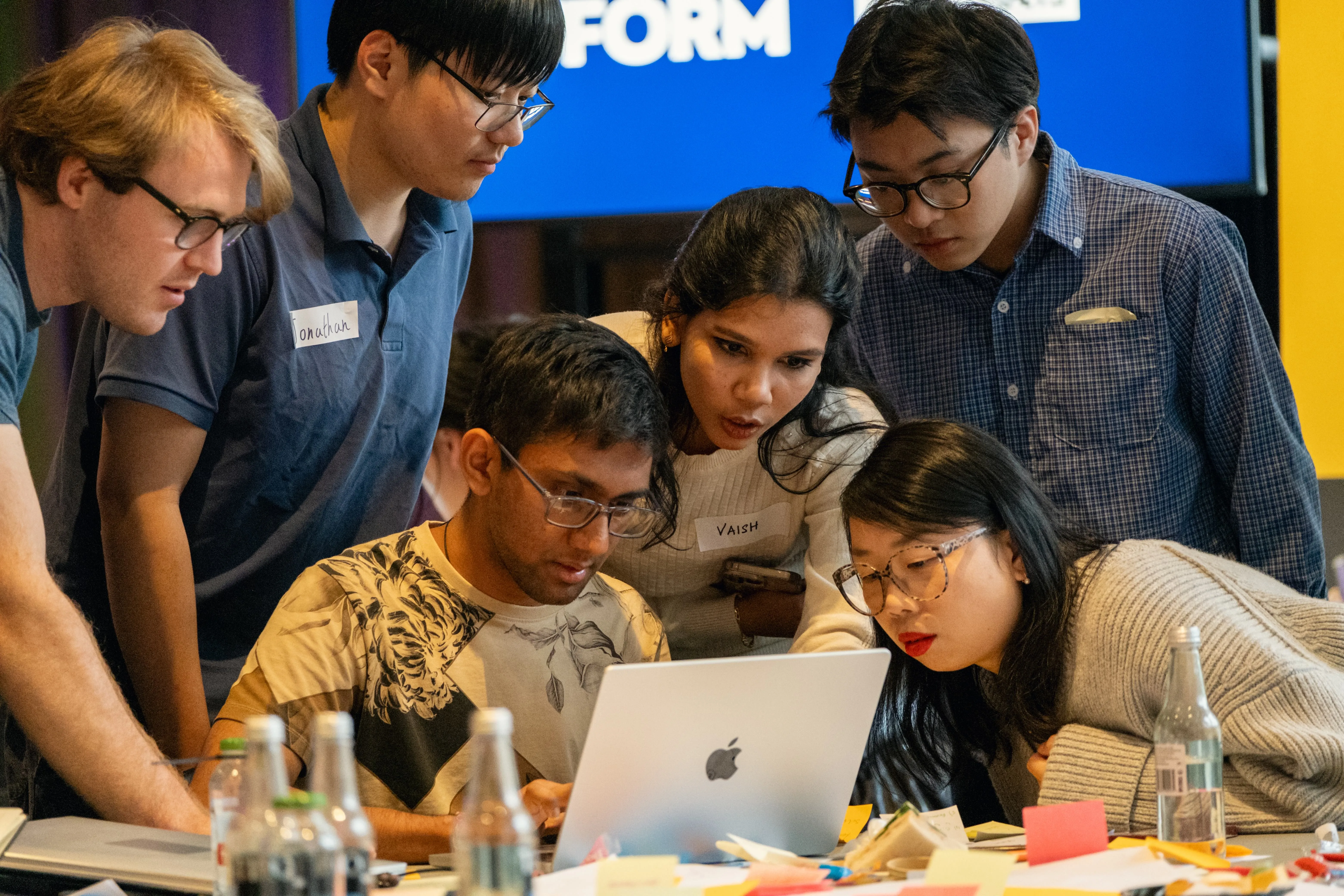 Six diverse young adults focused on a laptop screen, collaborating at a cluttered table with bottles and notes.