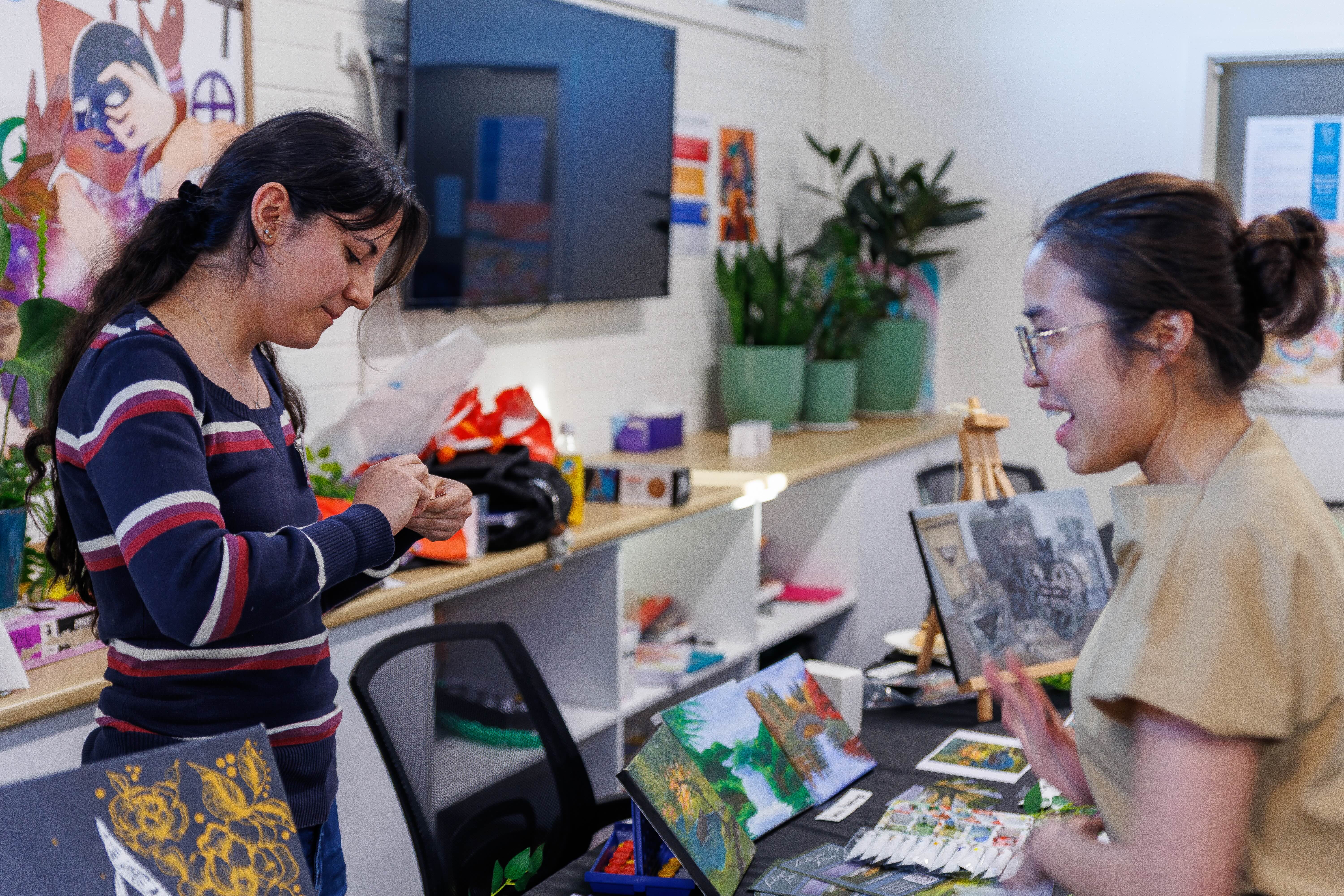 Two smiling young women with glasses standing close, one holding books, the other with headphones.