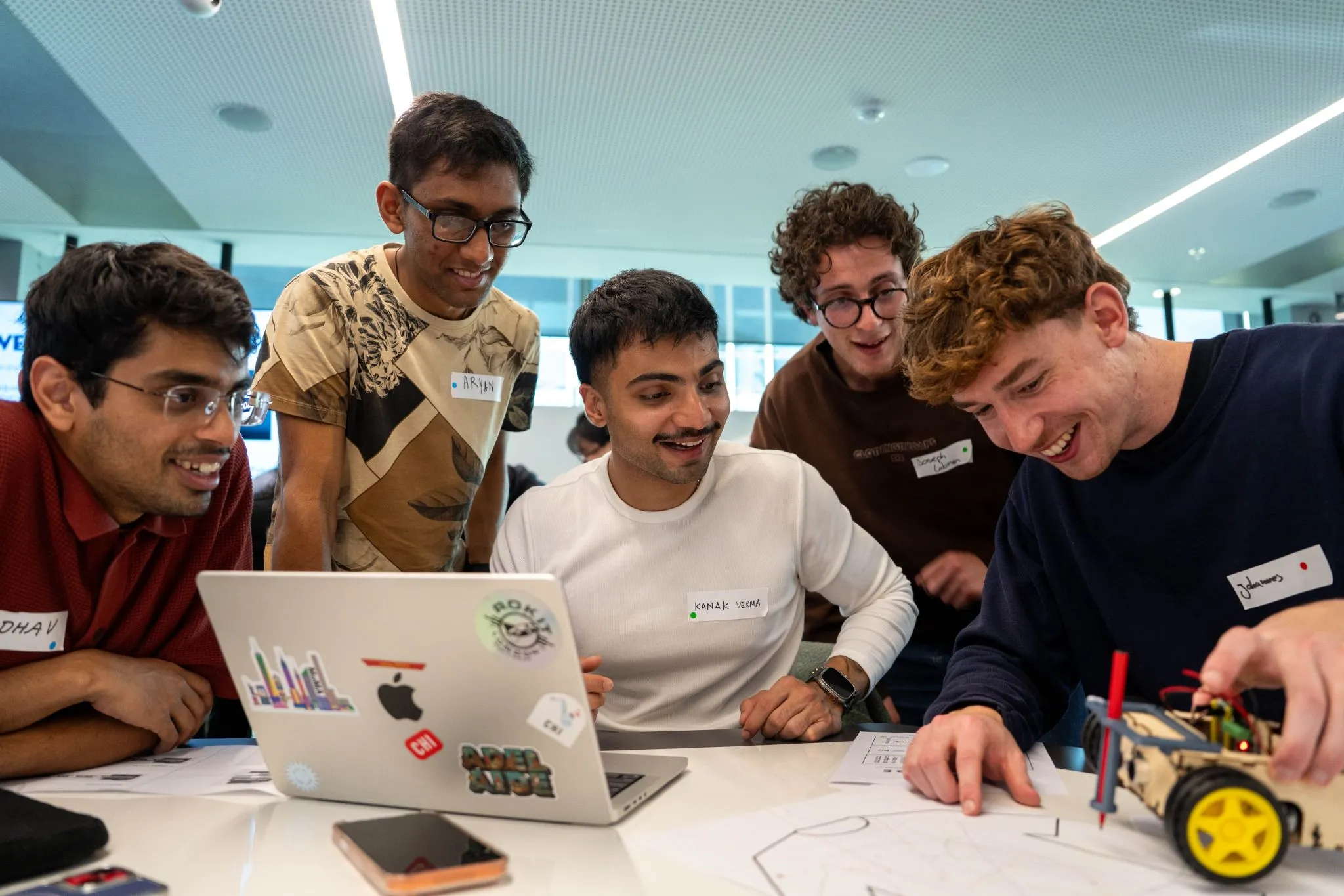 Five young men gathered around a laptop and small robot, working on a project together.