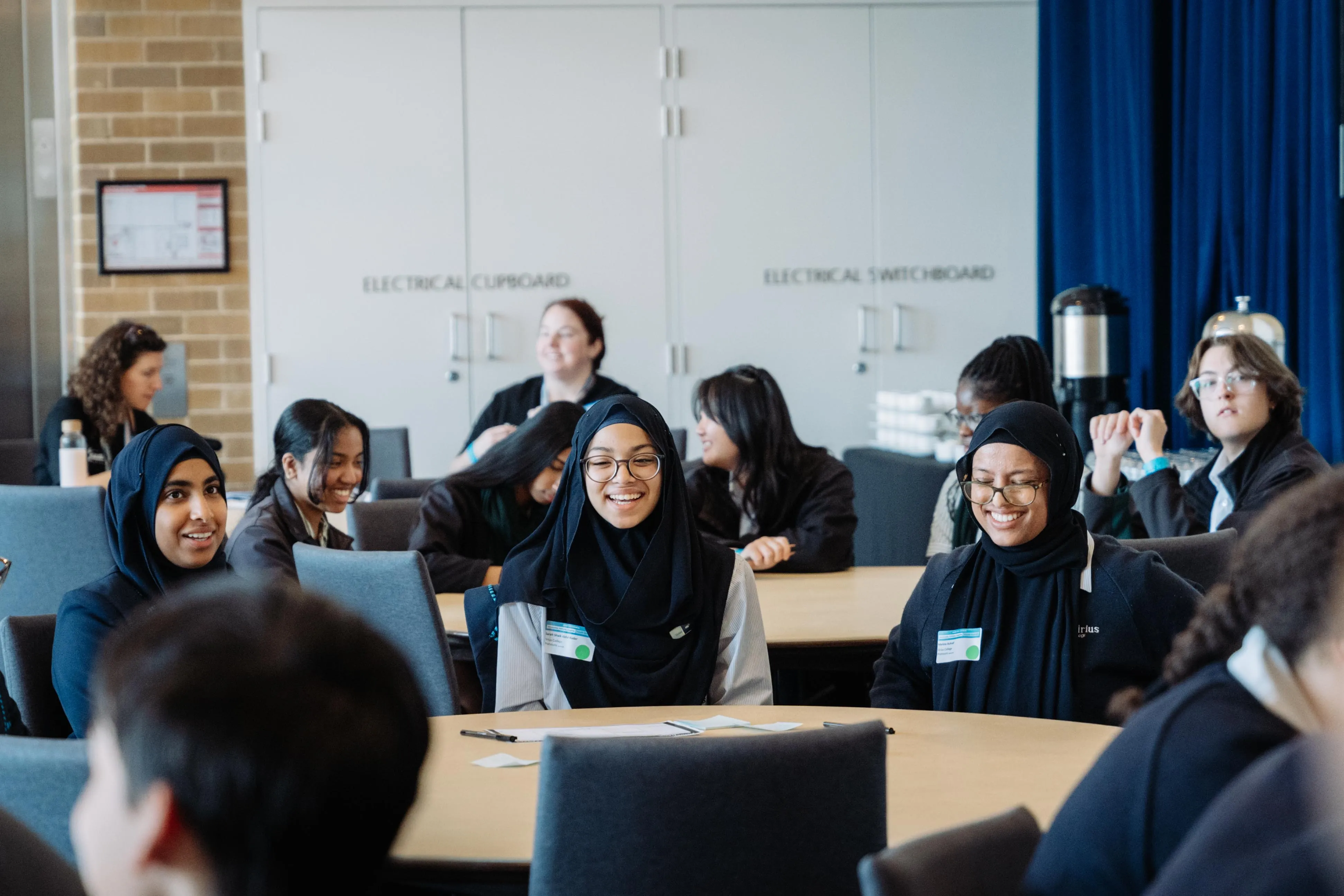 Group of young people sitting around a table, some wearing hijabs, smiling and engaging in conversation.