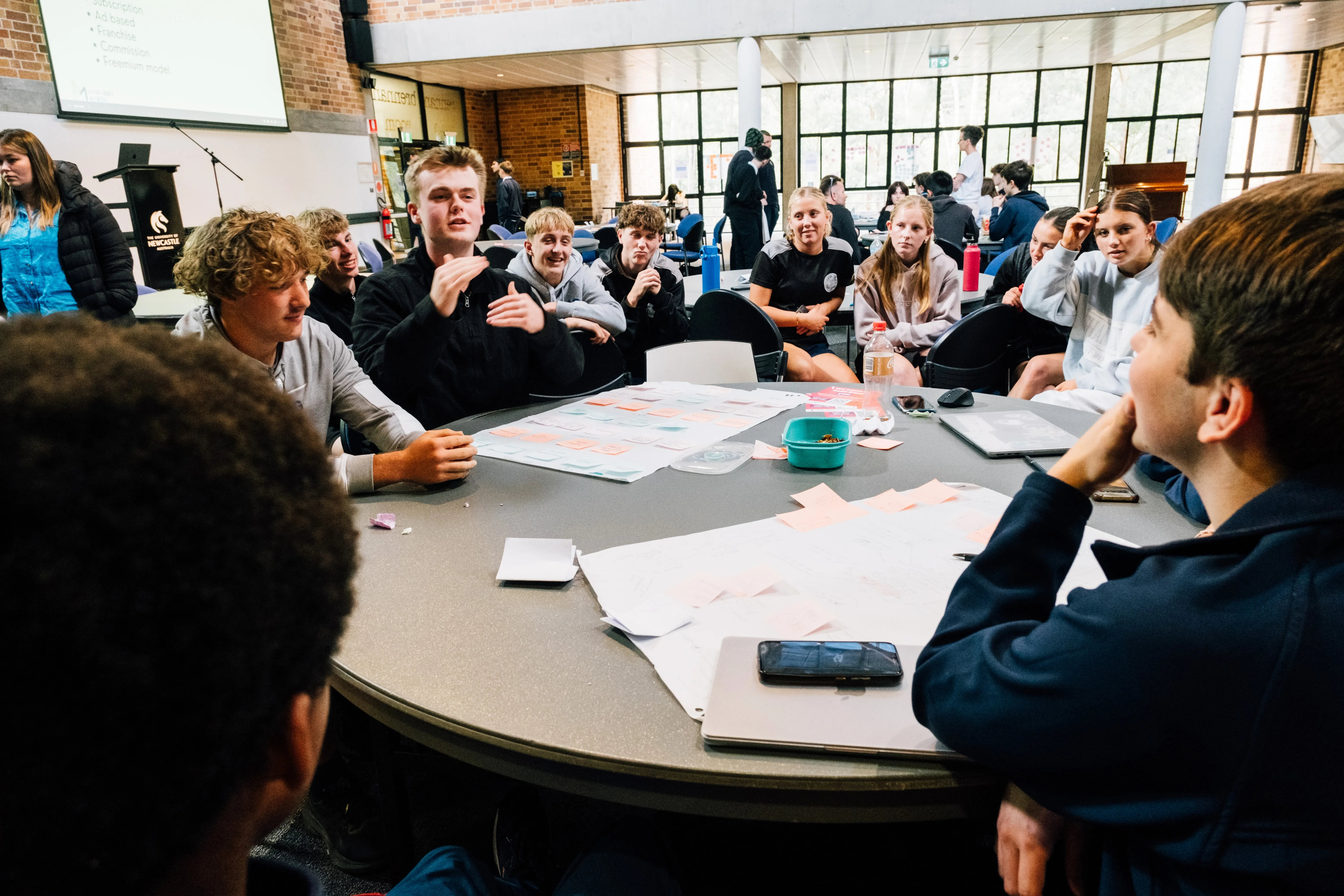 Group of students seated around a table engaged in discussion with notes and laptops in a classroom.