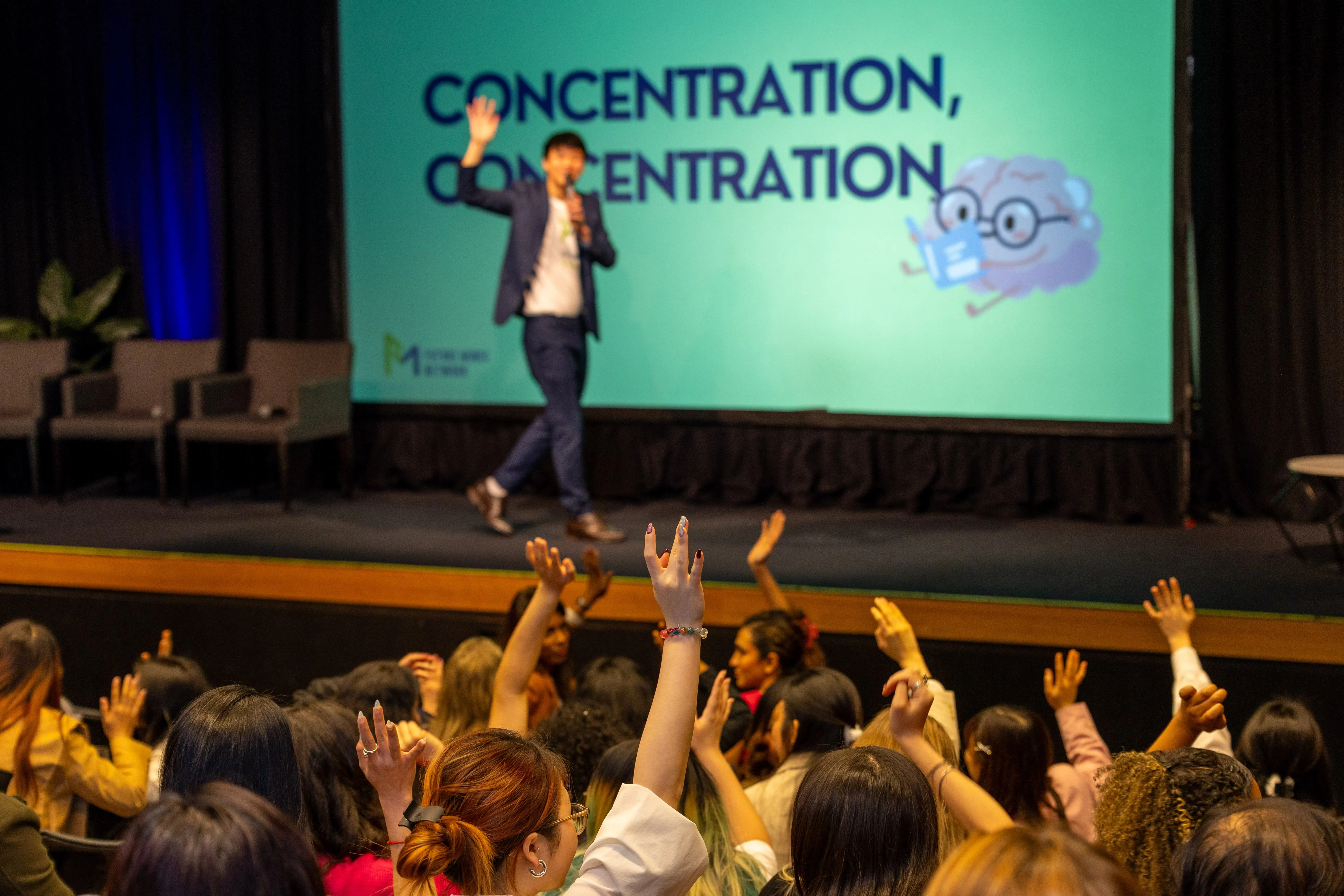 Audience with raised hands facing speaker on stage in front of screen reading 'Concentration, Concentration'.