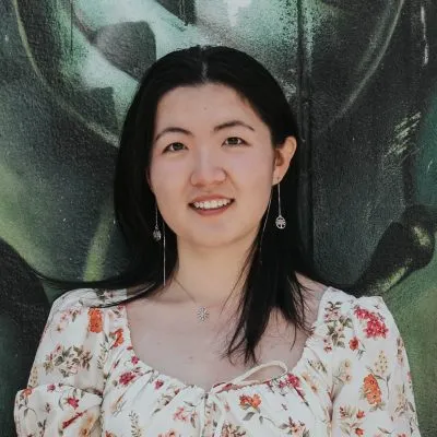 Smiling woman with long dark hair in floral blouse stands against graffiti wall background.