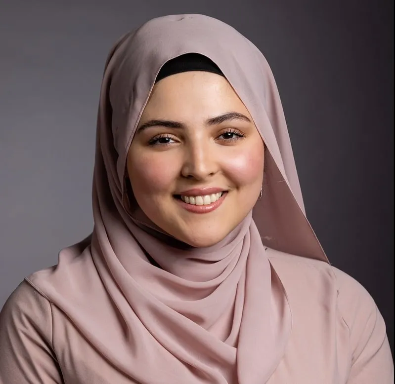 Smiling woman wearing light pink hijab and matching top against a gray background.