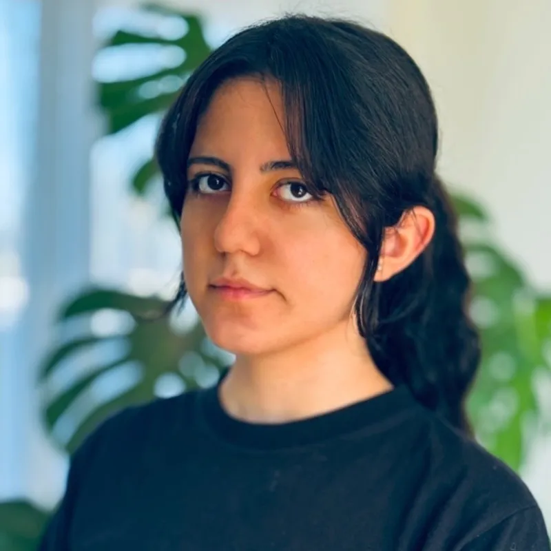 Young woman with dark hair in a ponytail wearing a black shirt, with blurred green plants in background.