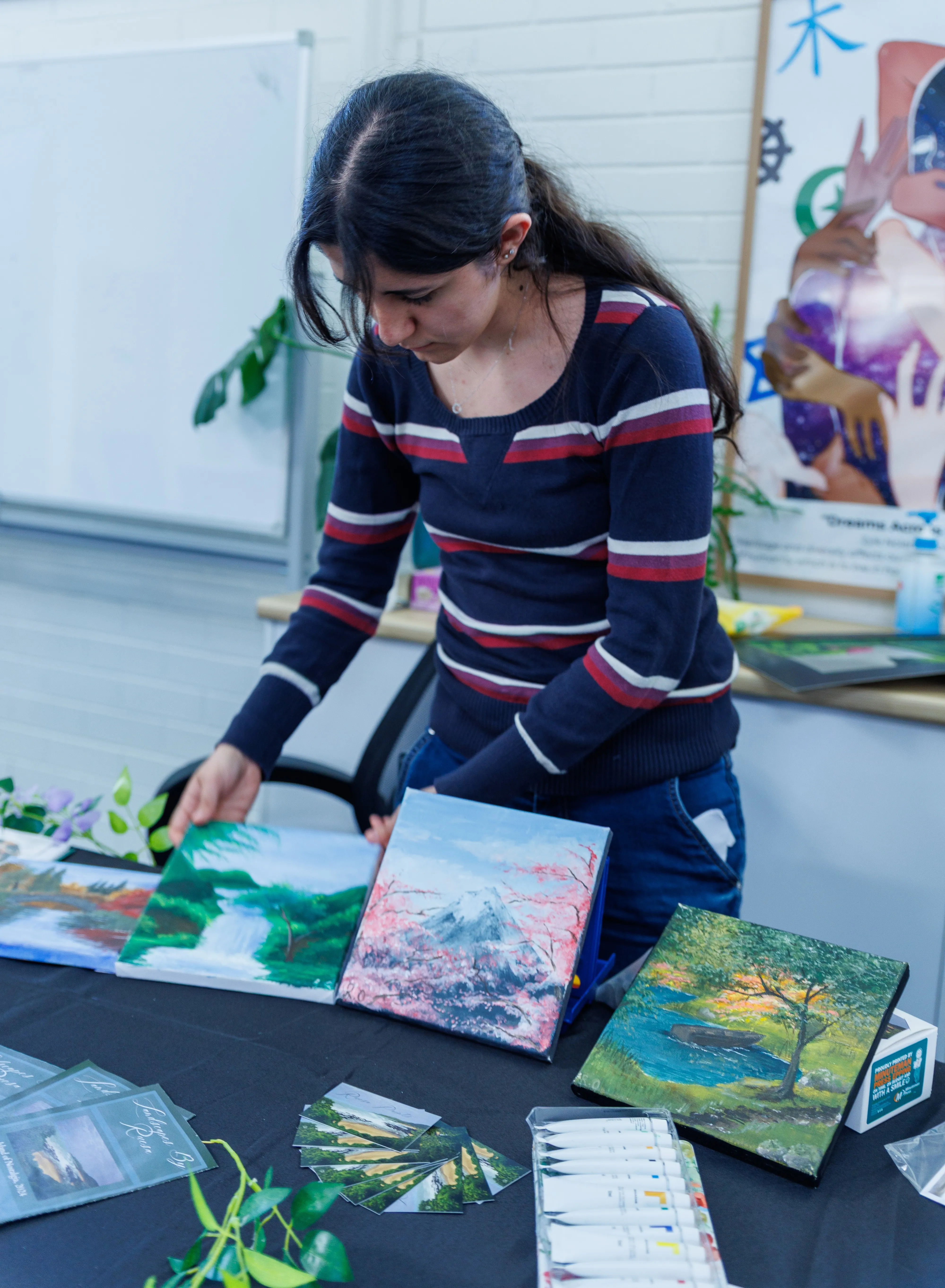 Woman arranging four landscape paintings on a table, with paint tubes and cards displayed.