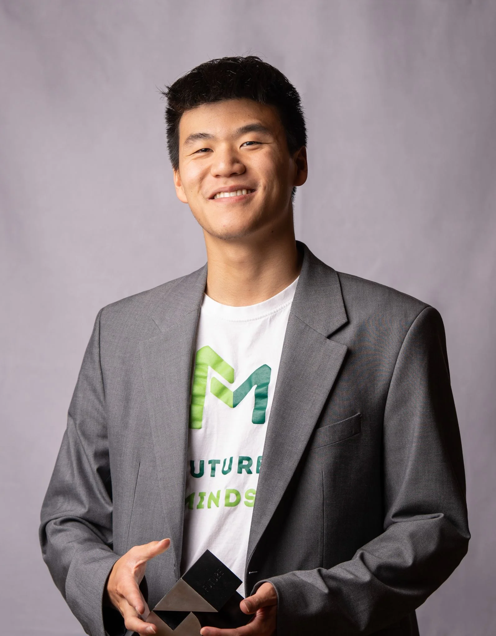 Young man in a gray blazer smiles while holding a black geometric award trophy.