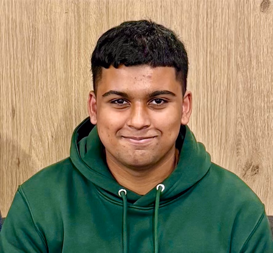 Young man with short black hair wearing a green hoodie against a wood-textured background.