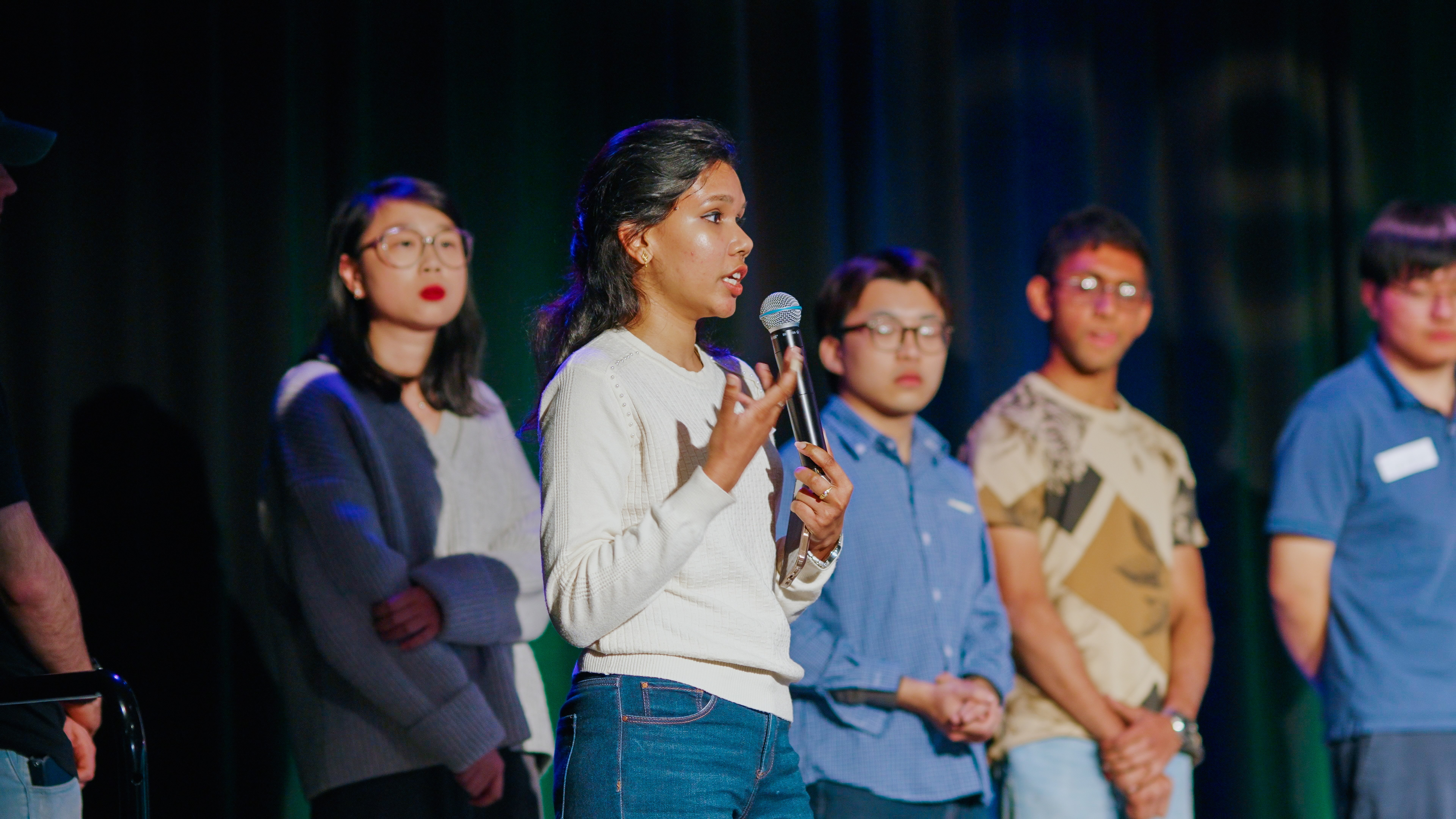 Young woman speaking into microphone with four people standing in line behind her in dim light.