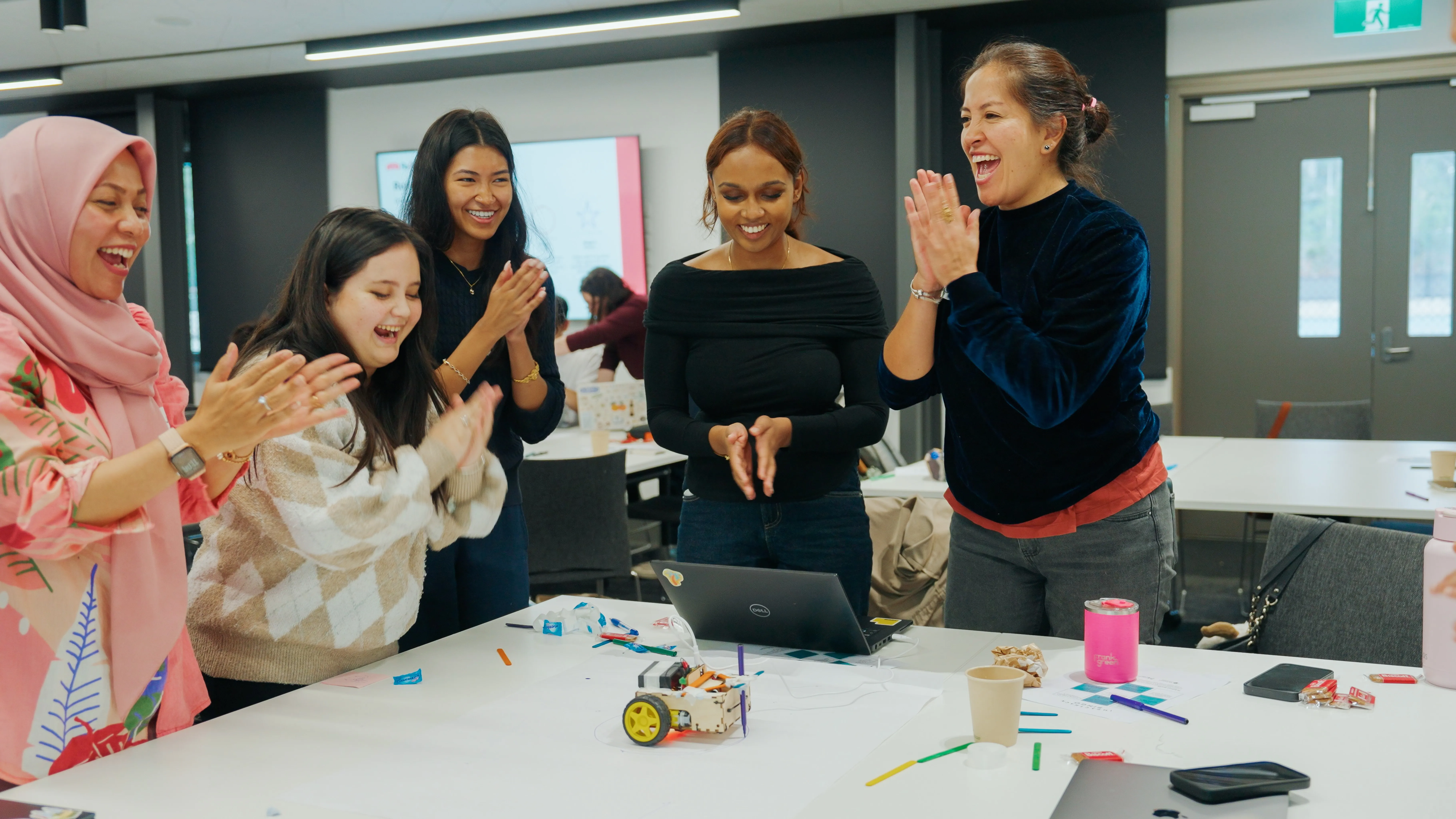 A diverse group of women clapping joyfully around a table with a small robot and laptop.