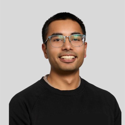 Smiling young man with black hair and glasses wearing a black sweater on a gray background.