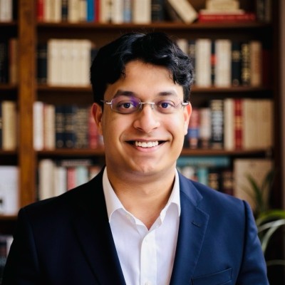 Smiling man with glasses wearing a dark suit and white shirt standing in front of a bookshelf.