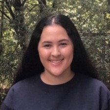 Smiling woman with long dark hair standing outdoors with trees in the background.