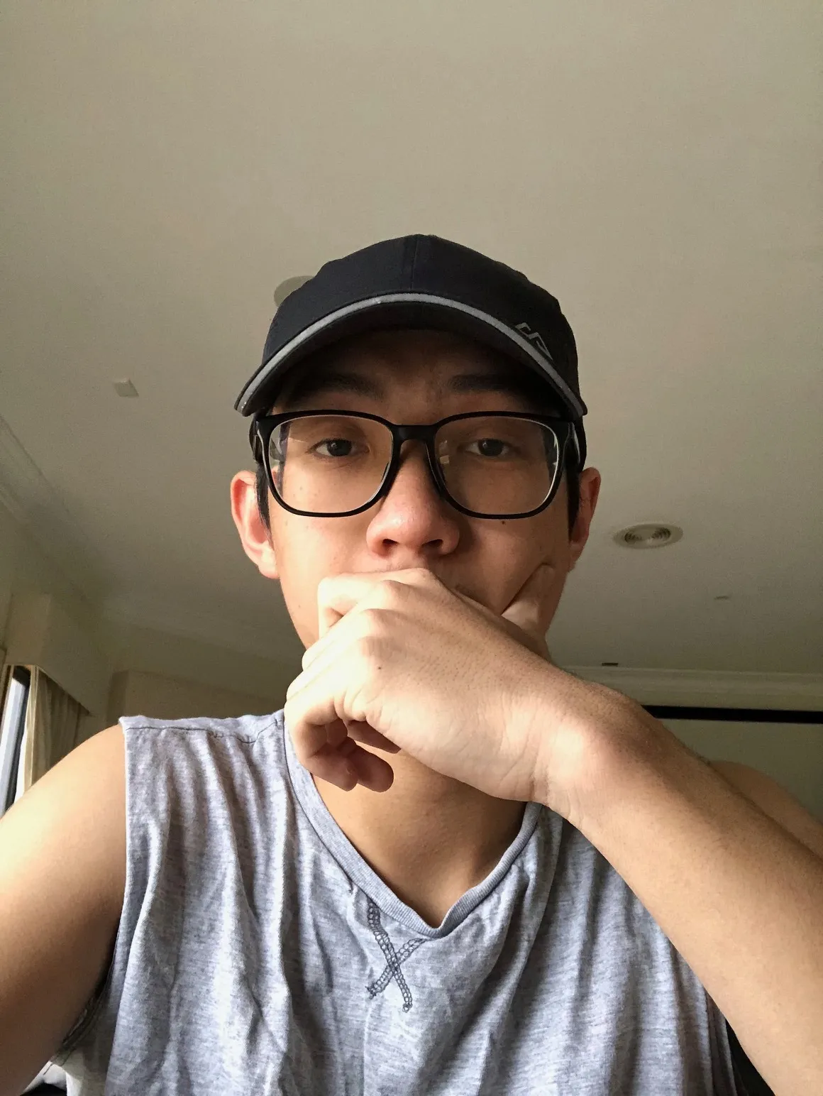 Young man wearing glasses and a black cap, resting his chin on his hand, indoors.