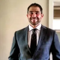 Smiling man in a dark suit and tie standing indoors against a light background.