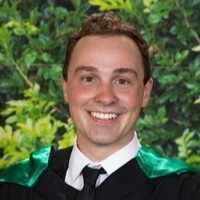 Smiling young man in a graduation gown and tie with green foliage background.