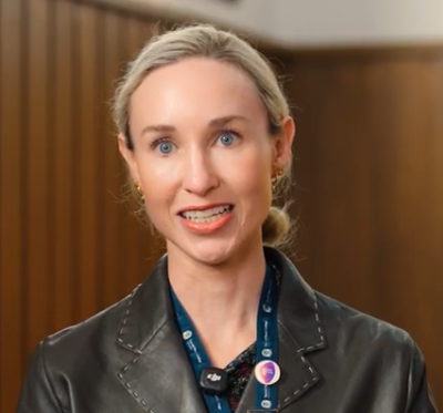 Woman with blonde hair tied back wearing a black jacket and a blue lanyard with pins.