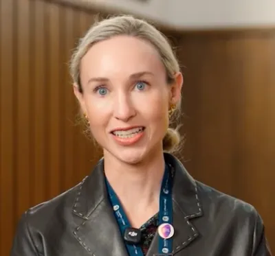 Woman with blonde hair tied back wearing a black jacket and a blue lanyard with pins.
