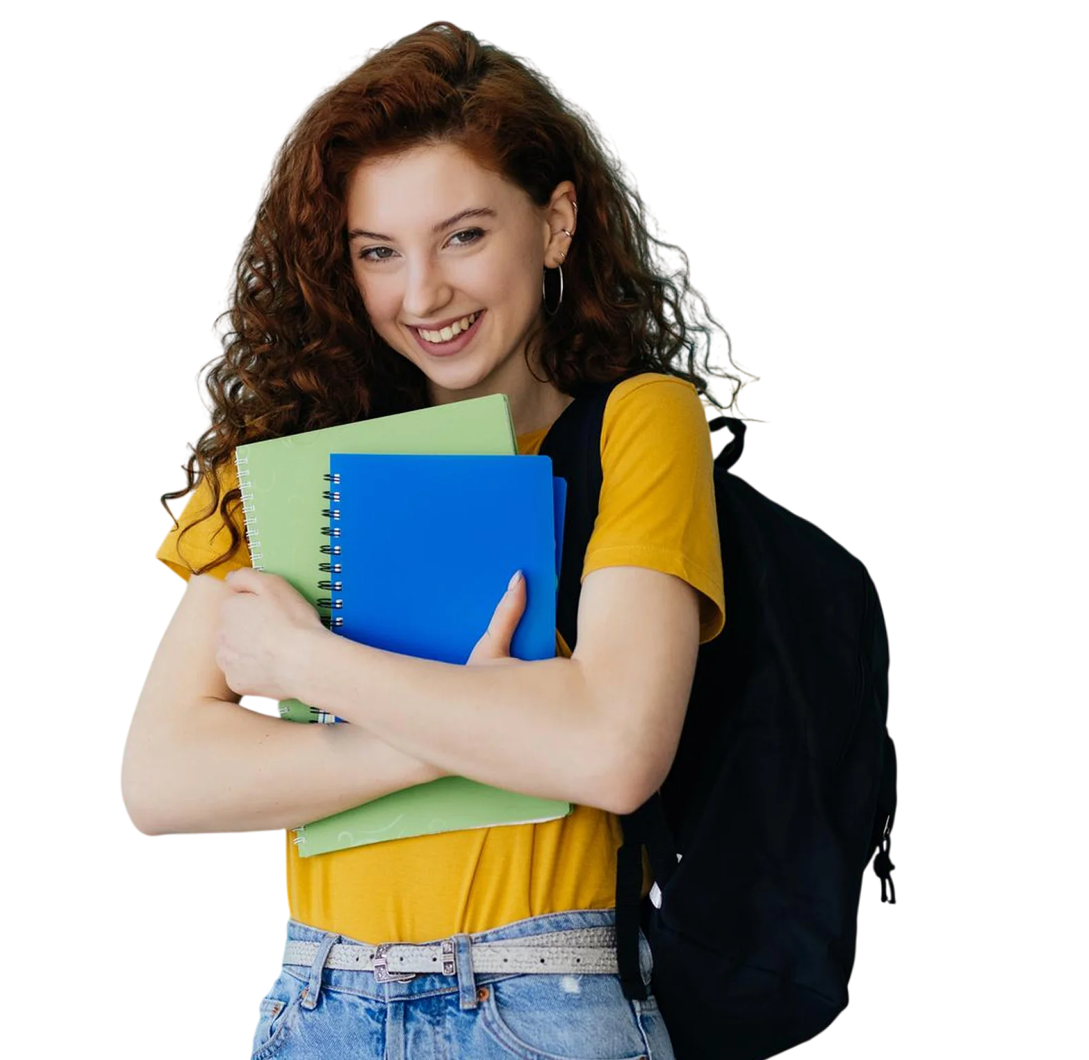 Smiling young woman with curly red hair wearing a yellow shirt and jeans, holding blue and green spiral notebooks and carrying a black backpack.
