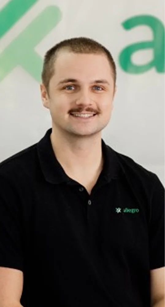 Smiling man with short hair and a mustache wearing a black polo shirt with a small green logo on the chest, standing in front of a blurred white background with green text.