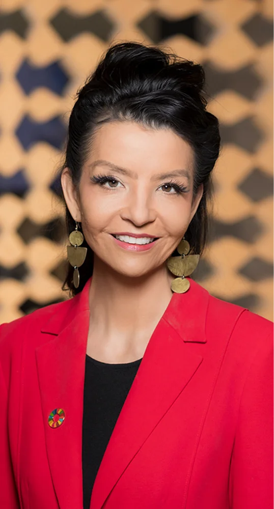 Smiling woman with dark hair styled up, wearing large gold geometric earrings and a red blazer over a black top, with a colorful circular pin on the blazer.