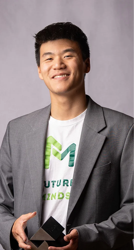 Young man with short black hair smiling, wearing a gray blazer over a white T-shirt with a green and blue 'M' logo and the words 'Future Minds', holding a geometric black and silver award against a plain gray background.