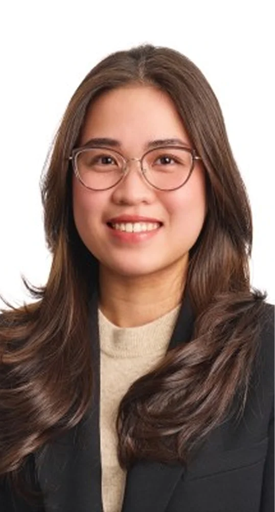 A young woman with long brown hair, wearing glasses and a black blazer over a beige top, smiling gently at the camera against a white background.