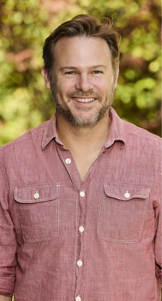 Smiling man with light beard wearing a faded red button-up shirt, standing outdoors in front of blurred greenery.