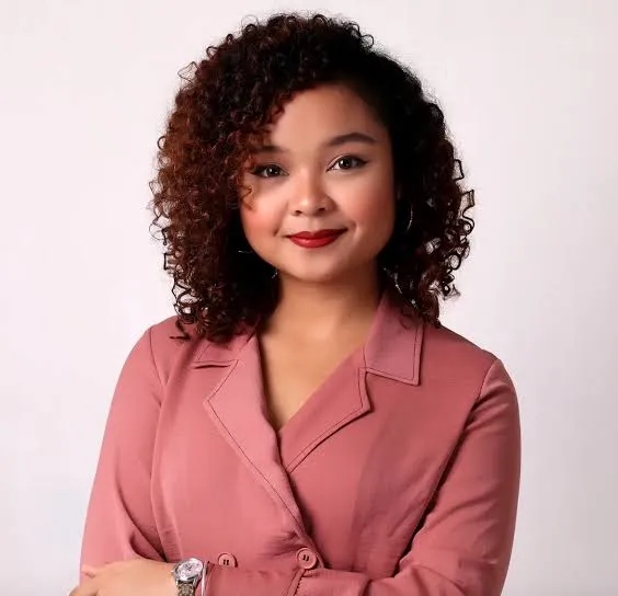 Woman with curly dark hair wearing a rose-colored blazer, smiling against a plain light background.