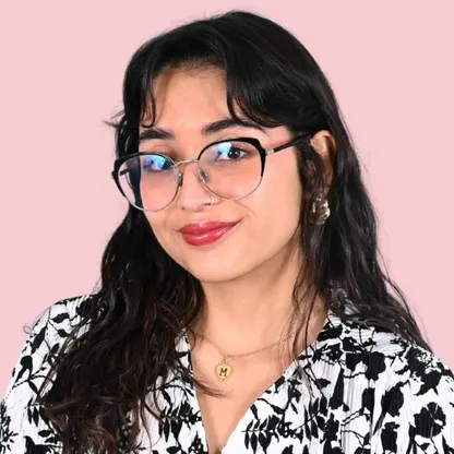 Young woman with long dark hair wearing large eyeglasses, red lipstick, and a black-and-white floral patterned top against a light pink background, smiling slightly.