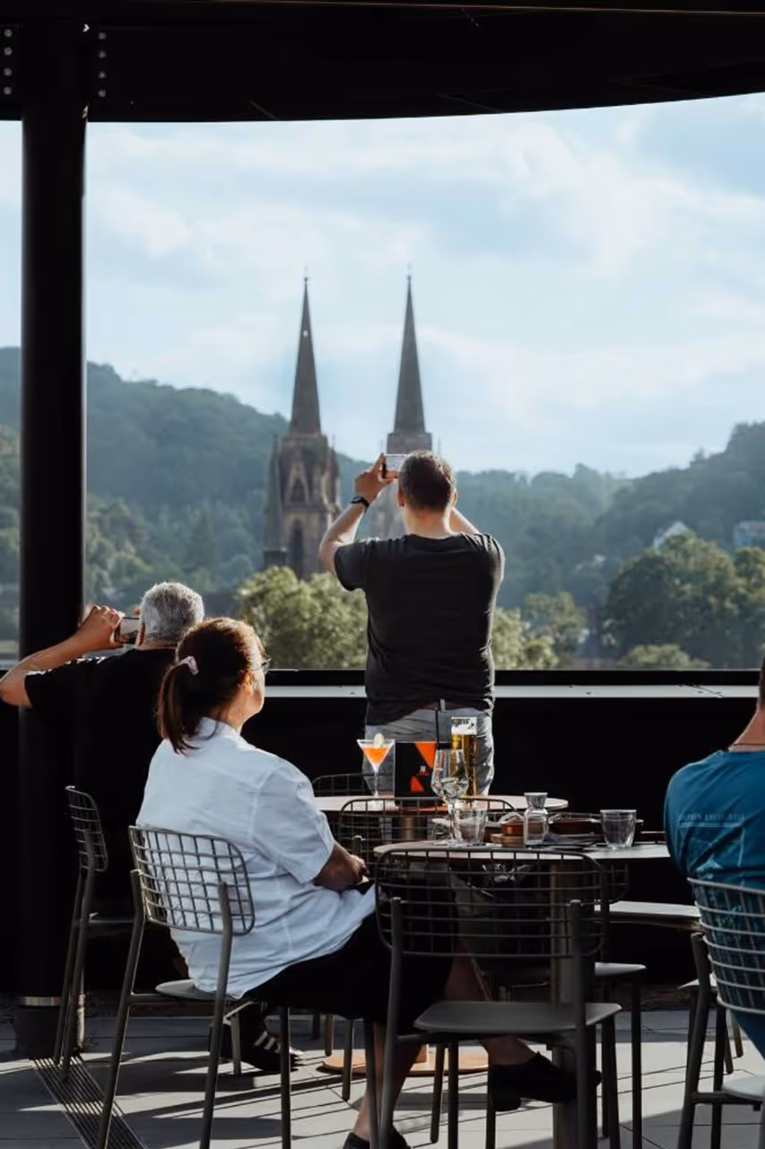People sitting and standing on a rooftop bar with drinks, overlooking church spires and trees in daylight.