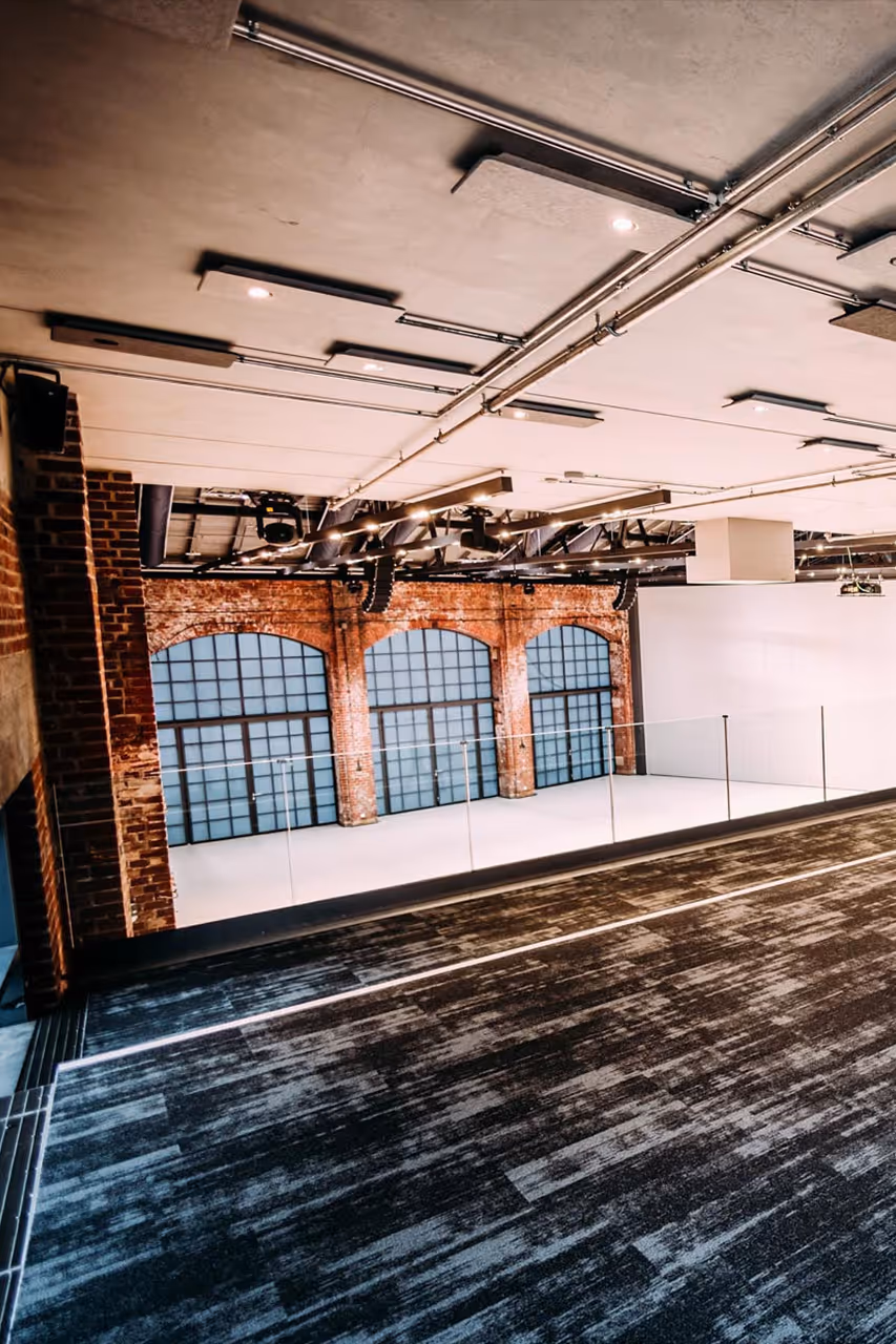 Modern auditorium with tiered dark flooring, brick wall, large arched glass doors, and industrial ceiling fixtures.