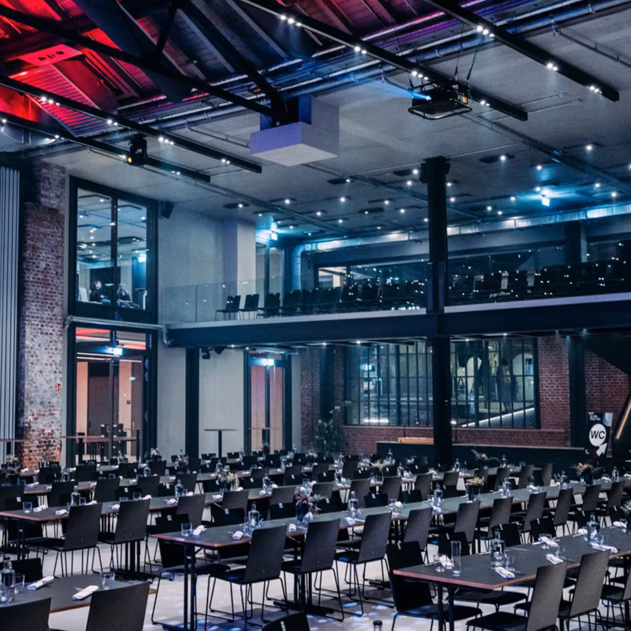 Modern industrial event space with rows of tables and chairs set for dining under exposed ceiling beams.