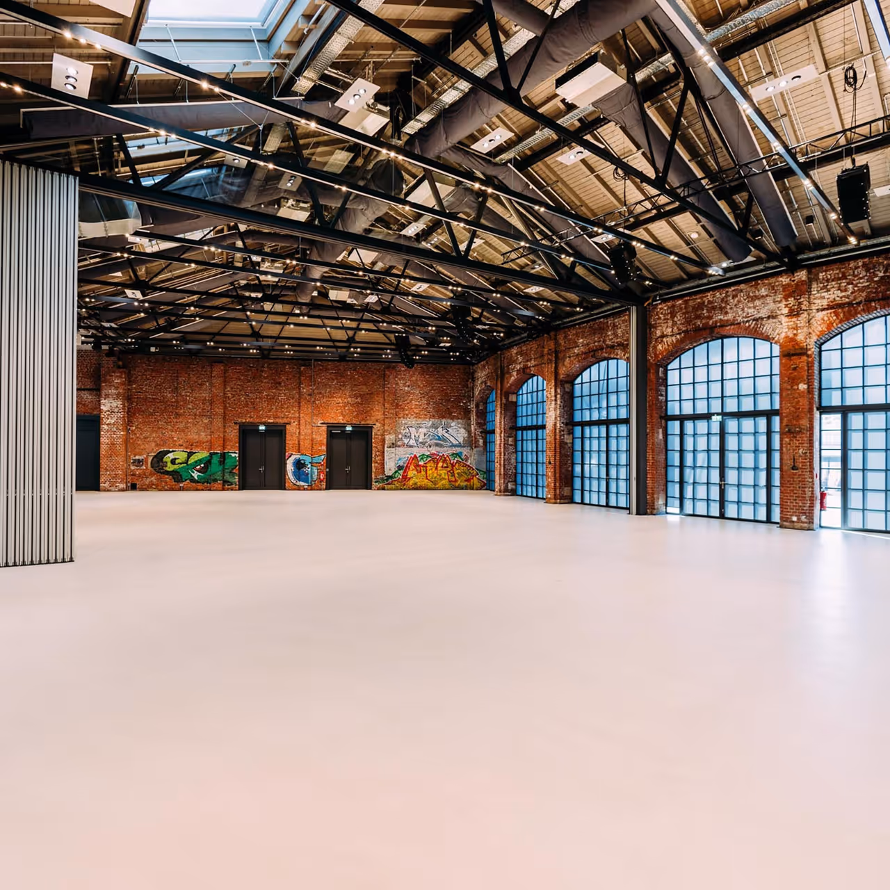 Empty industrial hall with brick walls, graffiti, large arched windows, and a high ceiling with exposed beams.