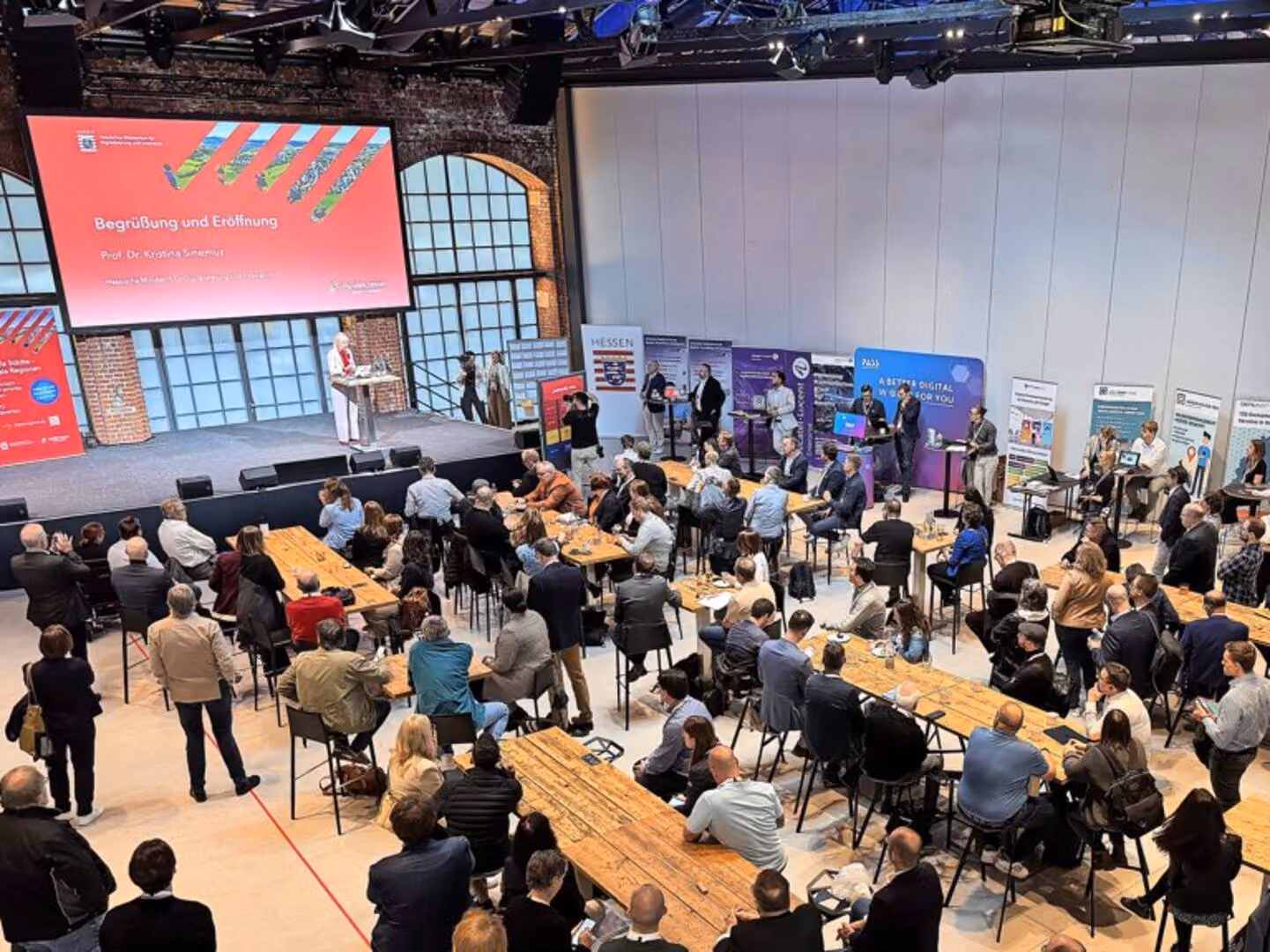 Audience seated at tables watching a speaker on a stage in a conference setting with banners behind.