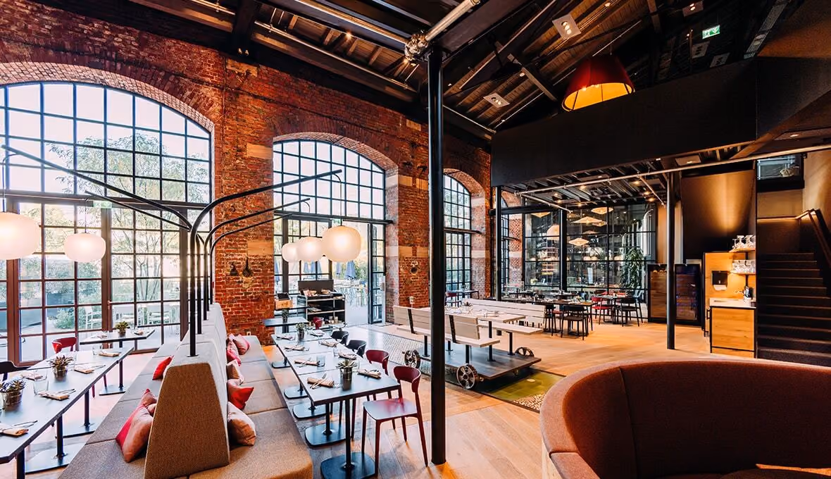 Spacious restaurant with large arched windows, brick walls, modern tables and chairs, and warm lighting.