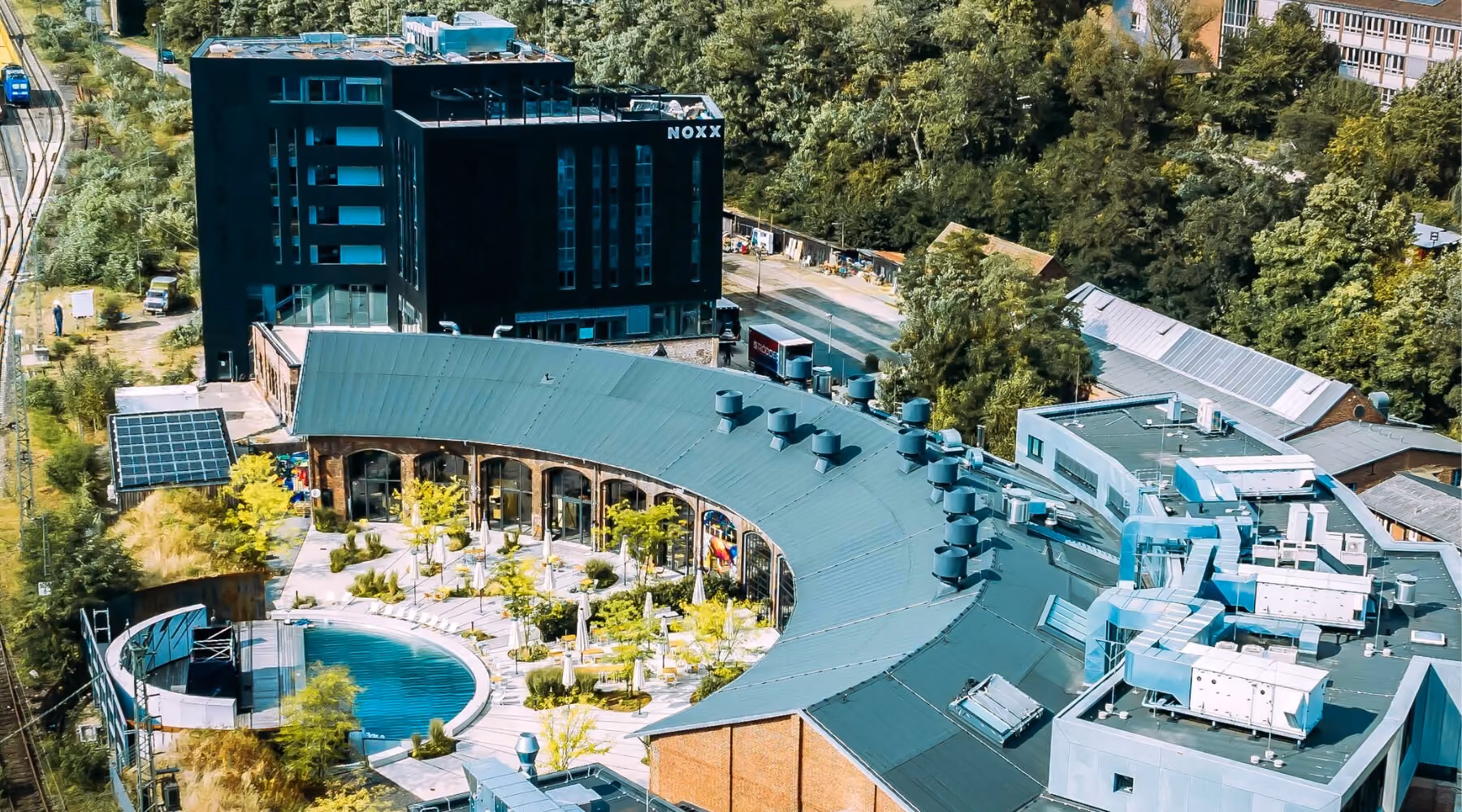 Aerial view of a curved building with outdoor seating and a round pool surrounded by trees and greenery.