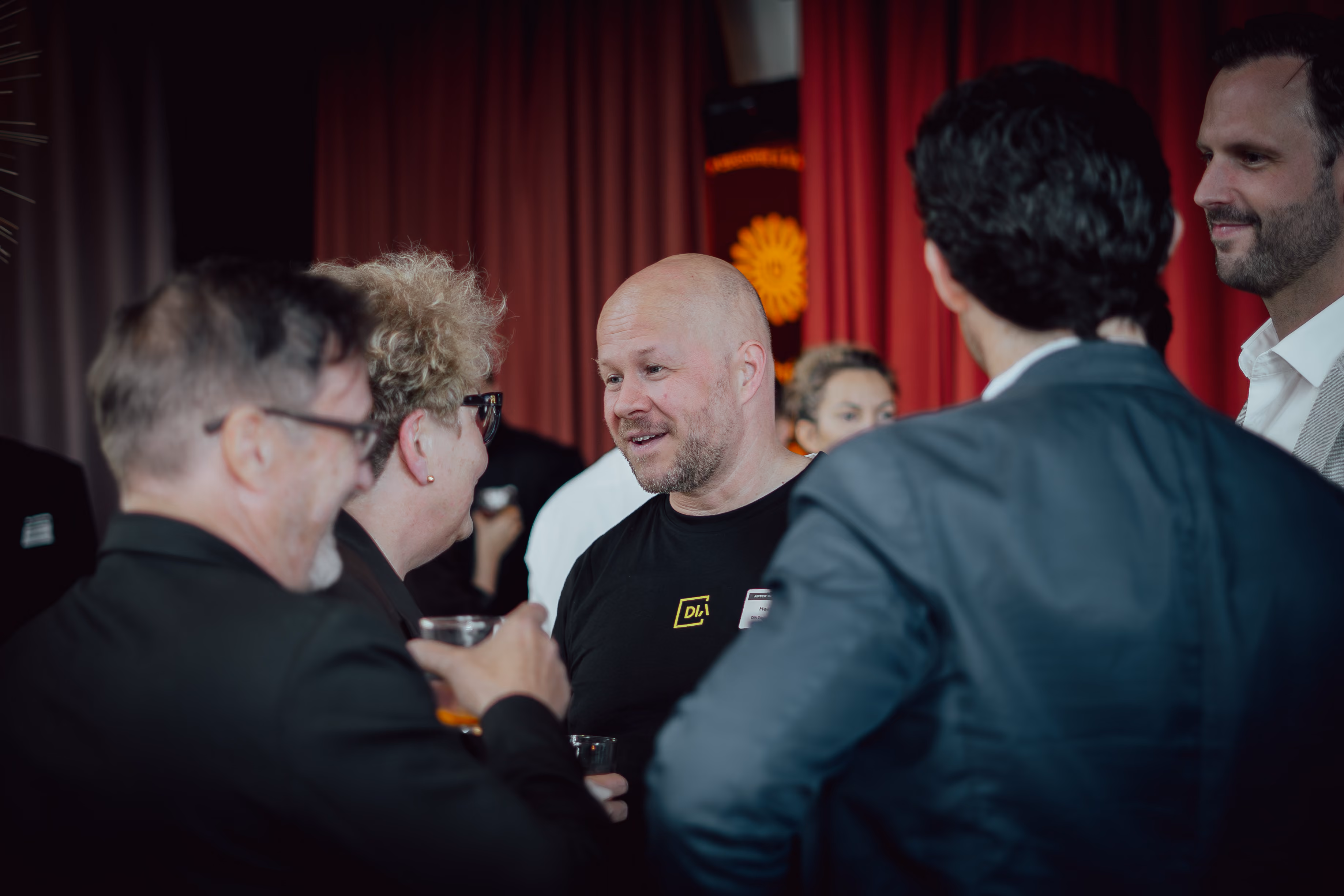 Group of people in conversation at an indoor event with red curtains in the background.