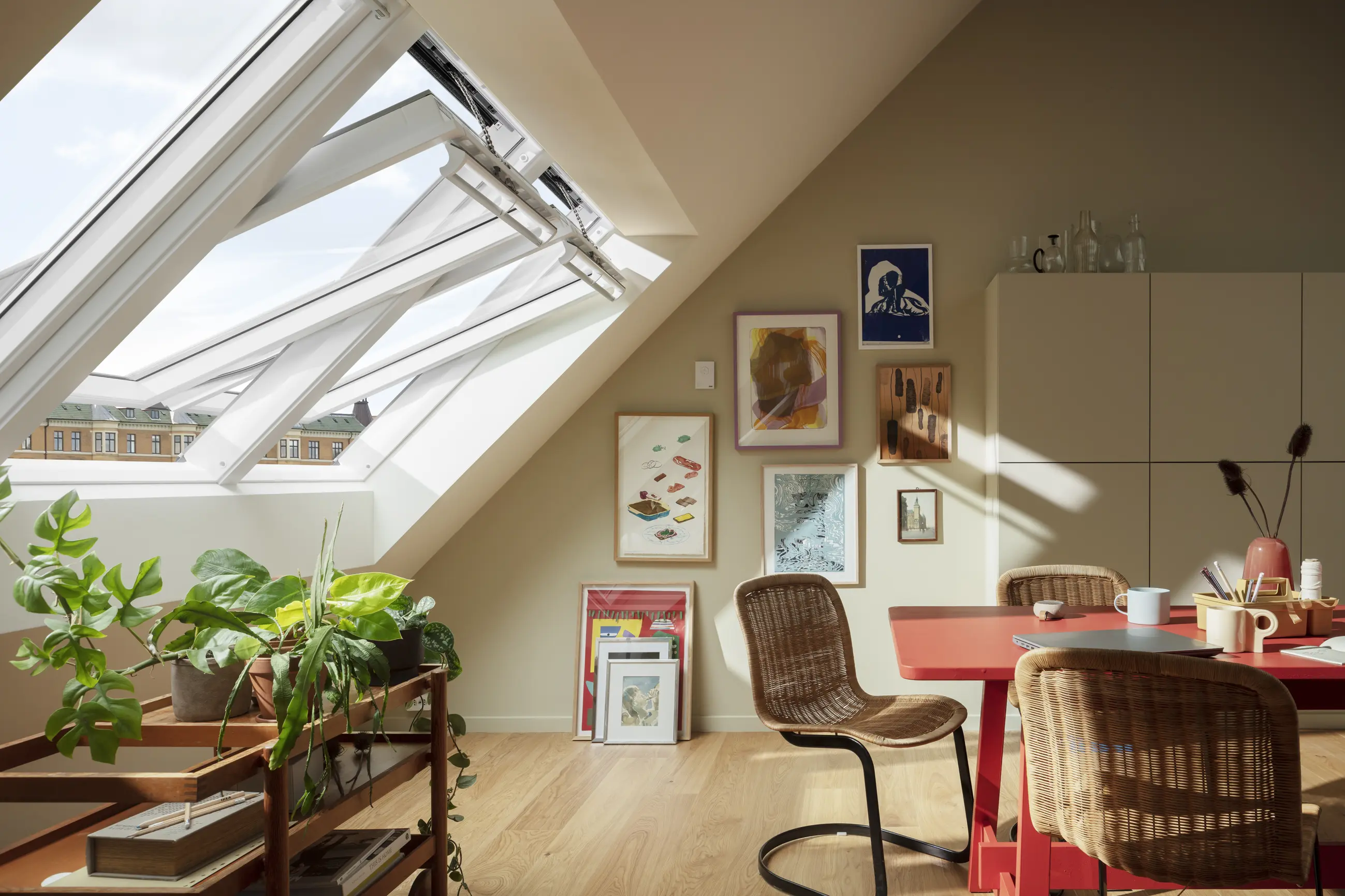 Helles Dachzimmer mit offenen Dachfenstern, Pflanzen auf einem Holzregal, rotem Tisch und Korbstühlen.