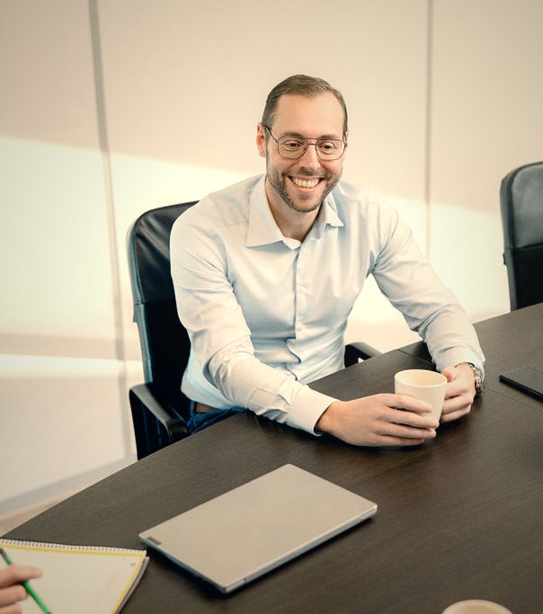 Lachende man met bril in wit overhemd zit aan een conferentietafel met een beker in zijn handen.
