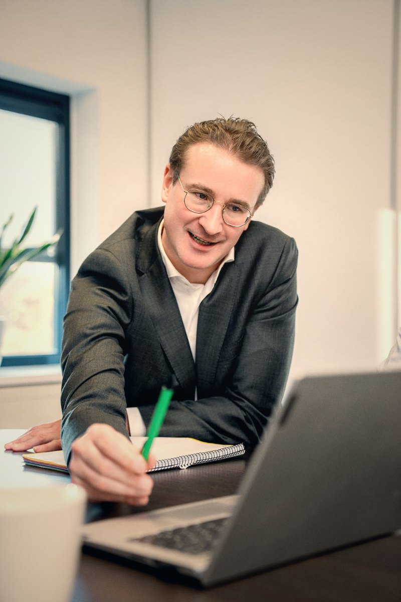 Zittende man in pak wijst met groene pen naar laptop terwijl hij glimlacht in een kantoor.