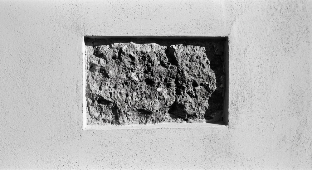 A smooth white rendered wall with a rectangular section cut away to leave one rough, weathered stone exposed in the middle